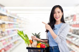 Beautiful Asian woman holding shopping basket full of vegetables and groceries in supermarket pointing to empty space aside