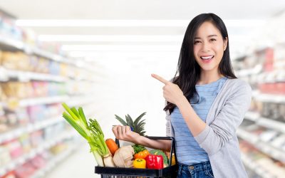 Beautiful Asian woman holding shopping basket full of vegetables and groceries in supermarket pointing to empty space aside